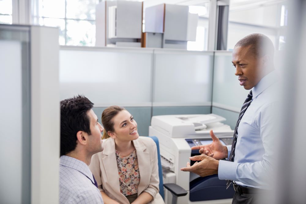 A businessman speaks with two professionals with a copier in the background signifying printer rentals