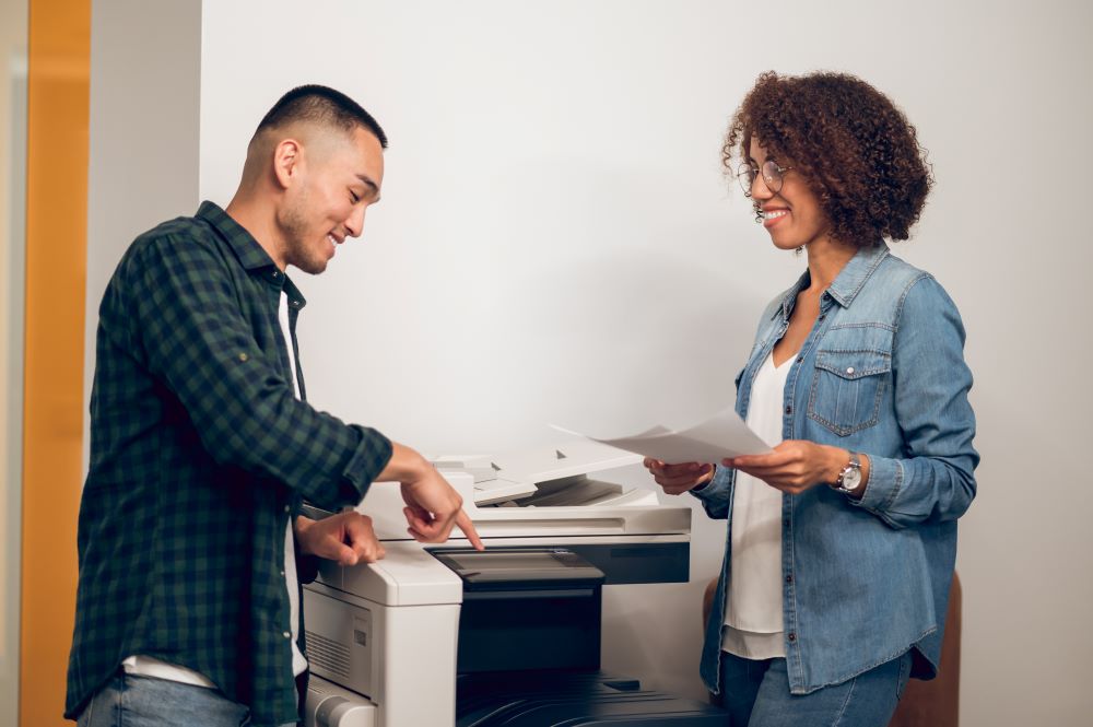 Two young coworkers gather and look at outputs at one of their office copiers.
