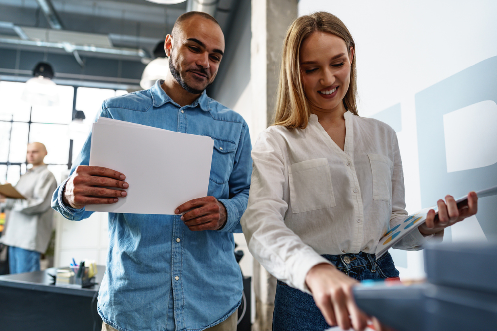 Two employees stand at a printer in an office holding dozens of printed documents symbolizing Managed Print Services.