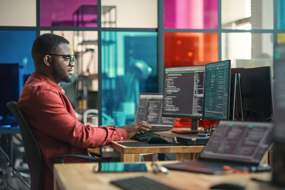 A young professional sits behind two monitors with code on the screens in a modern office signifying IT Services.